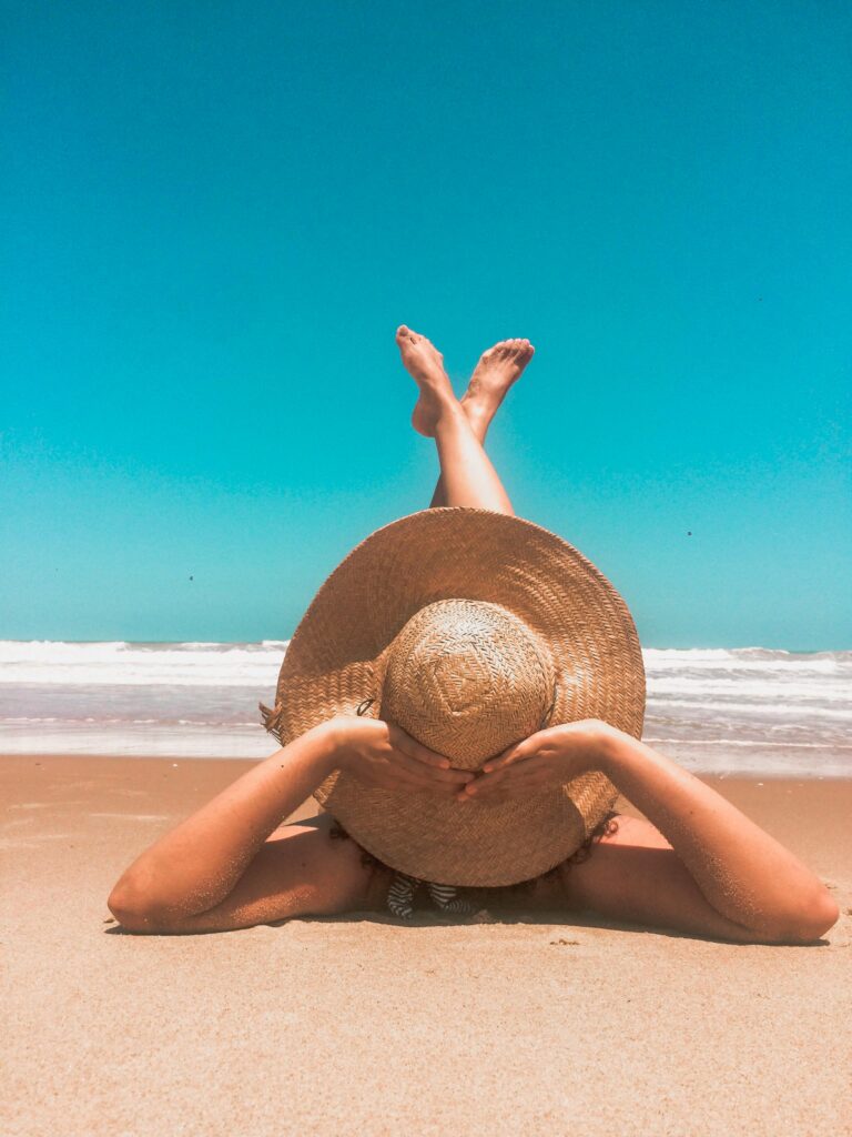 A woman in a straw hat relaxes on a sunny beach, enjoying the clear blue sky and ocean waves.