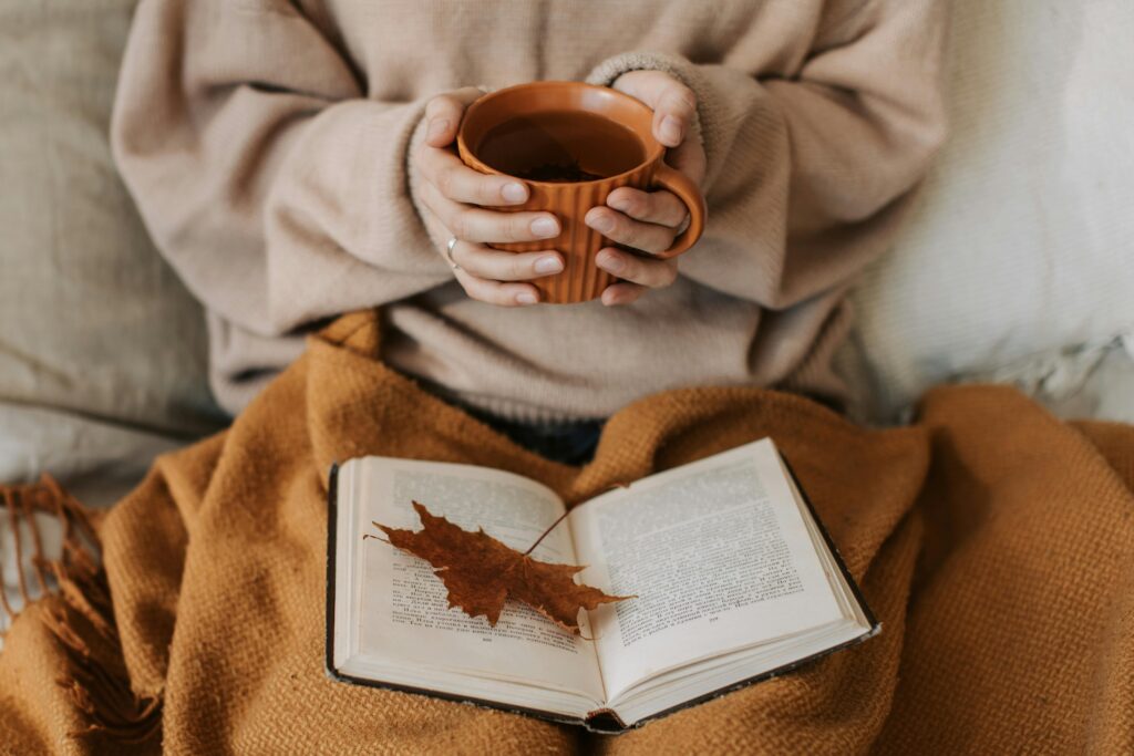 A woman enjoys a cozy autumn day, holding a tea mug over an open book with a maple leaf.